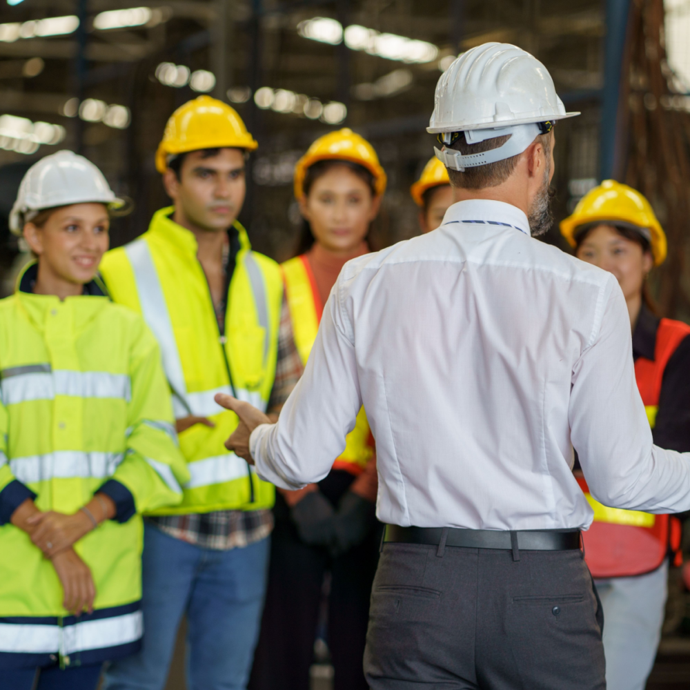 A diverse group of engineers of different genders and ethnic backgrounds stands together on-site at an industrial location. They are wearing hi-vis and hard hats for safety.