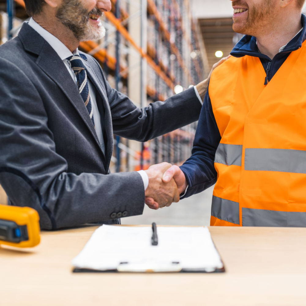 Two men shake hands in a warehouse, one wears a suit and the other wears a high-vis jacket.