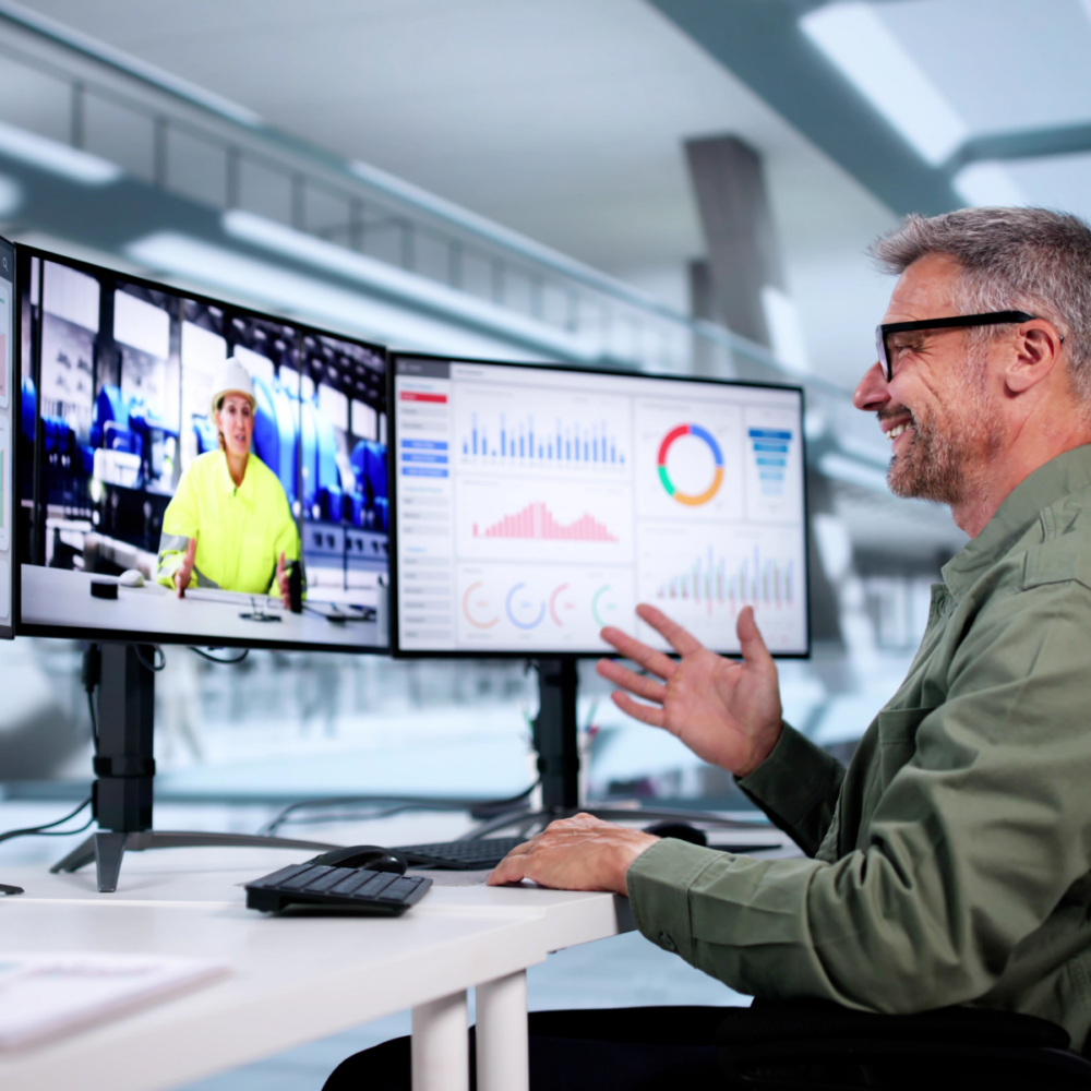 A man with grey hair wearing a green jacket is sitting at a desk in an office environment, conducting a video call with a female colleague on-screen who is wearing a hi-vis jacket and a hard hat on site.