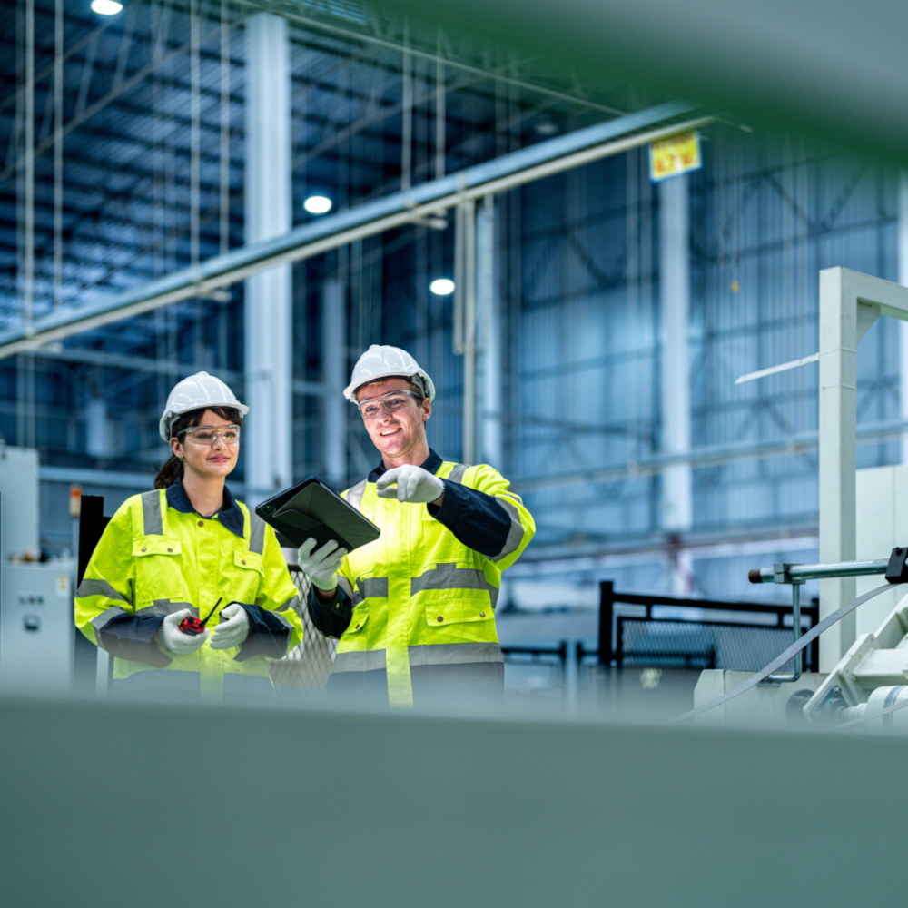 A male and female engineer stand in an industrial manufacturing site, both wearing high-vis yellow jackets and white hard hats. The man holds a digital tablet and the woman holds a walkie talkie.