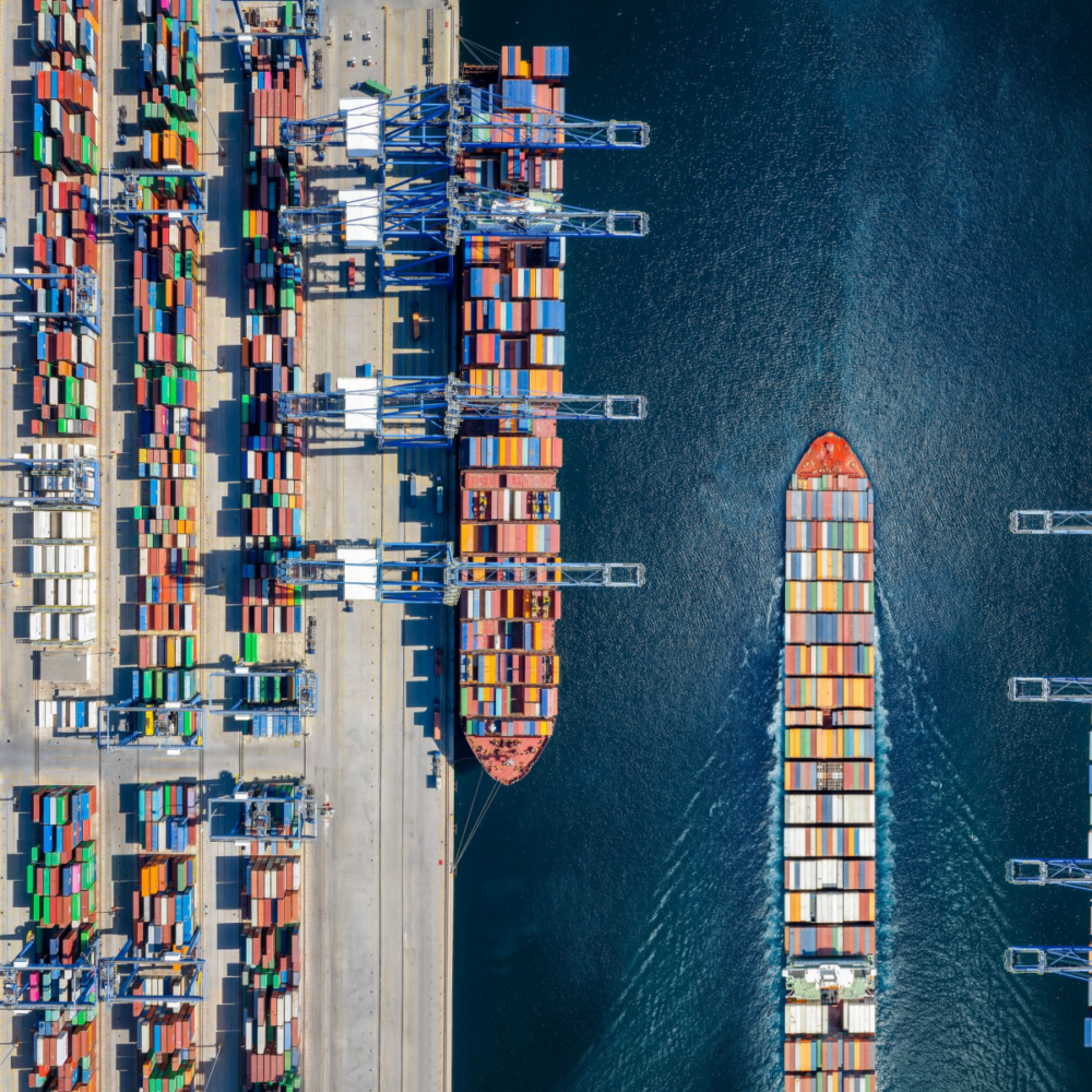 A port containing many shipping containers of different colours takes up the left third of the image. The middle third shows a container ship docked beneath a crane, in the process of being loaded. The right third shows a container ship sailing down a canal, loaded with containers.