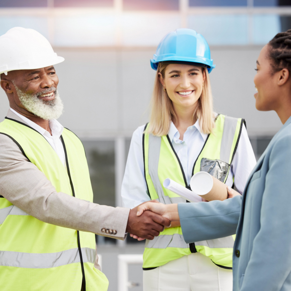 A black male engineer wearing a white hard hat and yellow high-visibility vest shakes the hand of a black woman wearing a pastel blue blazer. A white woman stands next to them, wearing a blue hard hat and yellow high-visibility vest and holding rolled-up engineering documents.